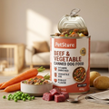 A can of PetStore Beef & Vegetable canned dog food stands open on a wooden table, surrounded by real food props including carrots, peas, potato, meat cubes, and a bowl in a softly blurred living room setting.